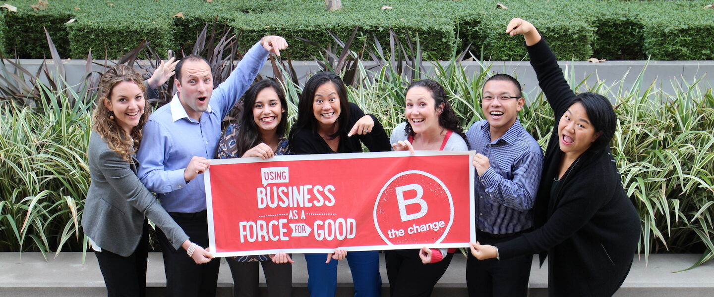Blue Garnet team members celebrate becomine a B Corp as they point to a red and white banner that says 