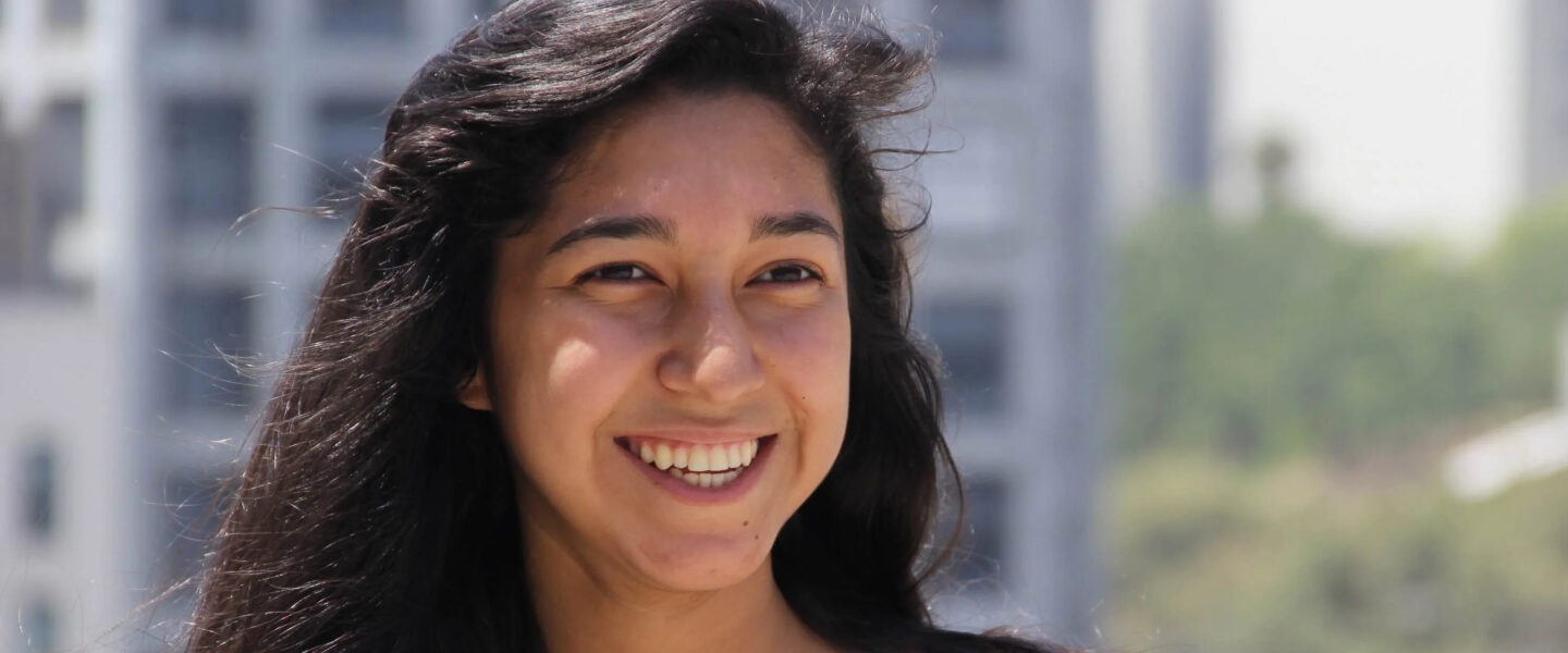 A young woman with long brown hair smiling in the sun, with a city view in the background