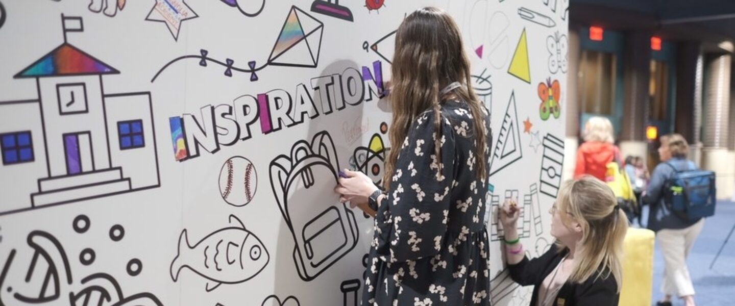 A woman with wavy brown hair stands in front of a wall with outlines of shapes on it. She is coloring in the word 