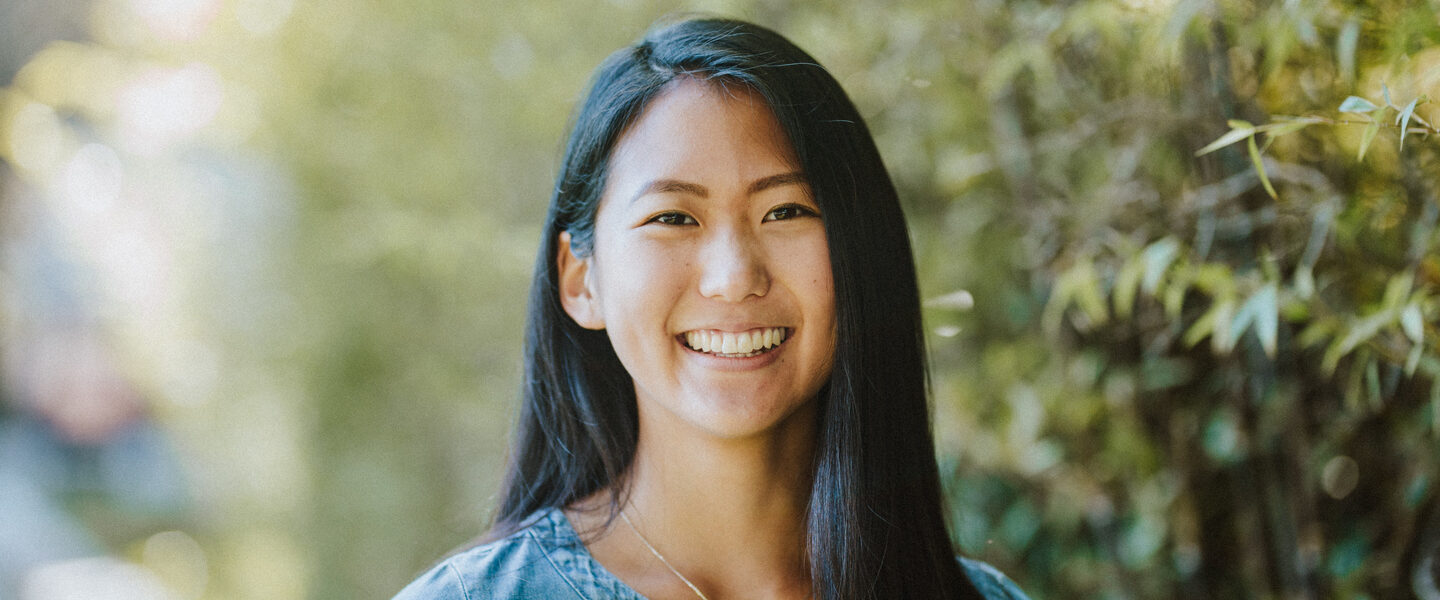 An Asian woman with black hair and wearing a blue blouse stands against backdrop of trees