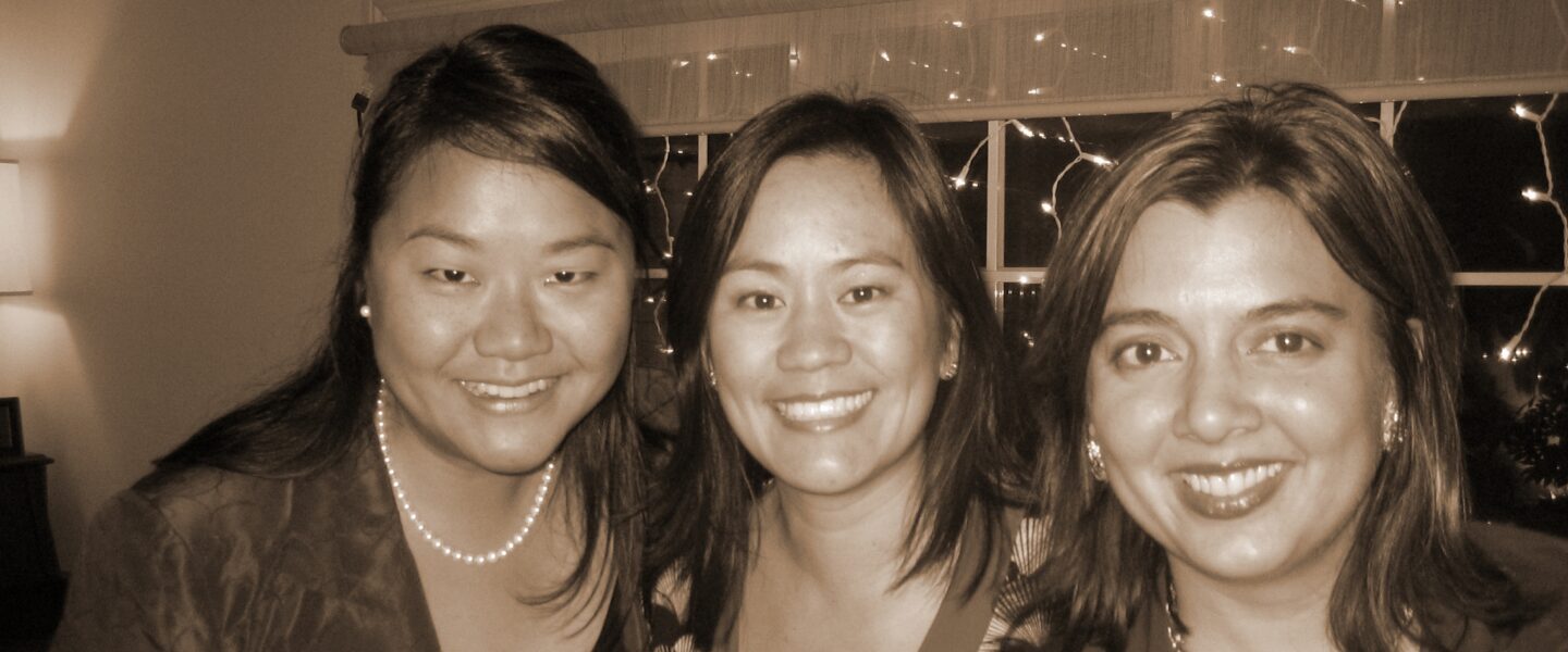 Three women stand in front of a window with Christmas lights in the background