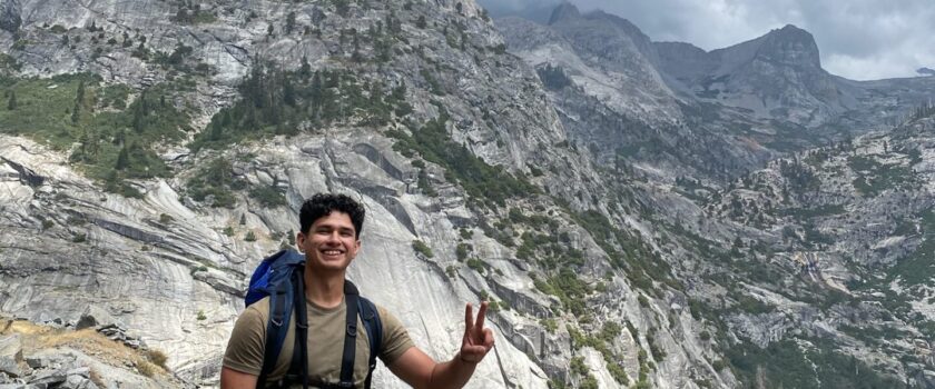 A man with brown hair smiles as he stands in front of a mountain