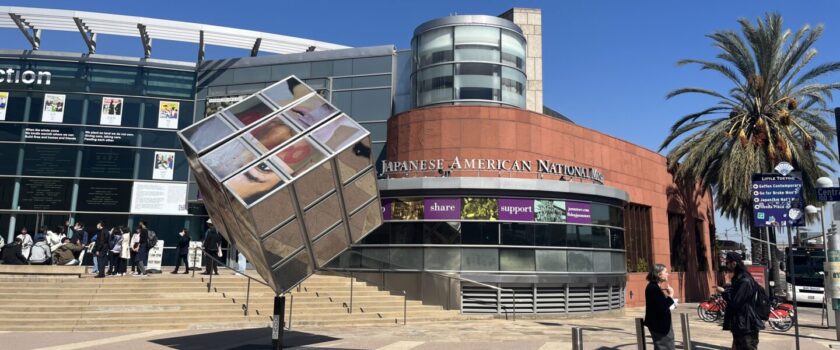 Two people stand and talk in front of the museum, a large glass building inset into red brick. There is a cube-shaped sculpture next them, with photos representing different aspects of human identity on each cube face.
